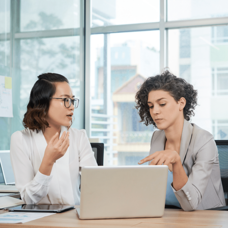 two woman sitting at a desk reviewing items on a laptop screen