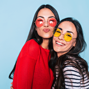 two brunette women smiling wearing colorful glasses against a blue backdrop