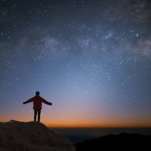 person in a red jacket with their arms outstretched on a rocky cliff against a blue starry sky
