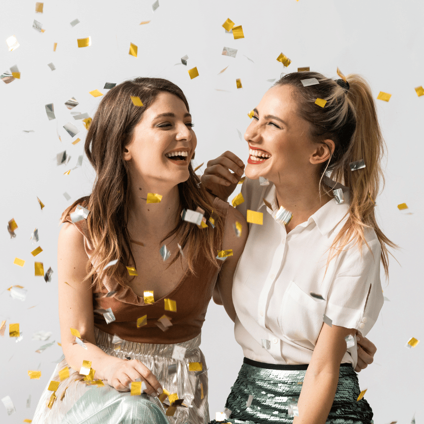two women looking at each other and smiling as confetti falls around them