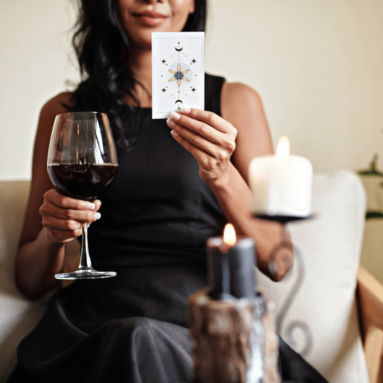 woman holding up a white tarot cared with black stars and moons and a wine glass filled with red wine behind a black and white candle