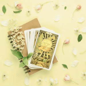 pile of white and yellow tarot cards on top of a notebook on a yellow table covered in pink and white flowers