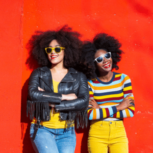 two women with afros in colorful clothing standing against a red backdrop wearing sunglasses smiling boldly