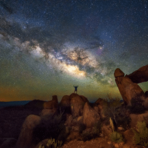 silhouette of a person standing atop a cluster of brown rocks with their arms outstretched to a blue and yellow starry sky