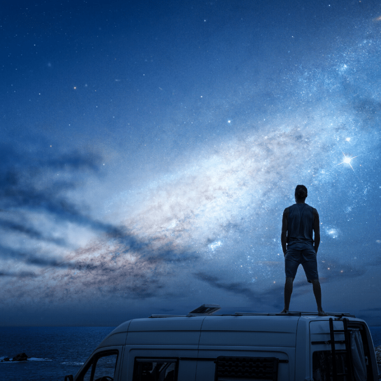 silhouette of a man standing on a camper van starring up at a blue starry sky