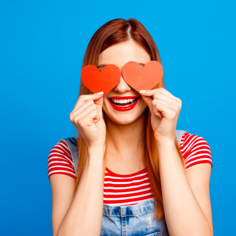 smiling redheaded woman in a striped shirt and blue overalls holding two red hearts up against a blue background