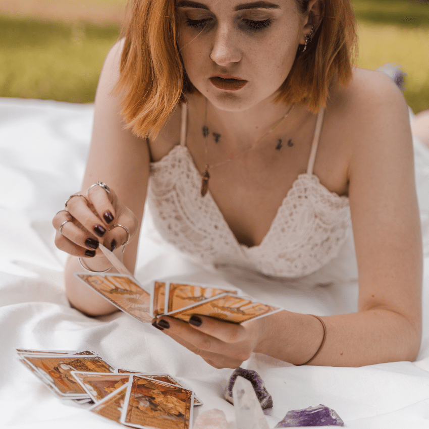 woman in a white shirt with short red hair laying on a white blanket reading tarot cards