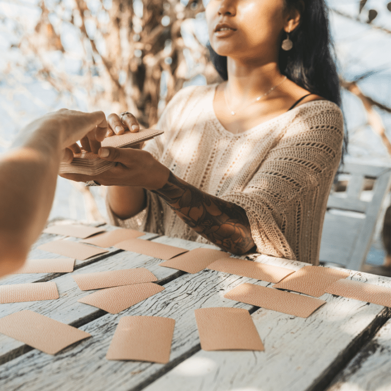 woman sitting at a wooden table covered in brown tarot cards holding out a deck of tarot cards for another person to choose from