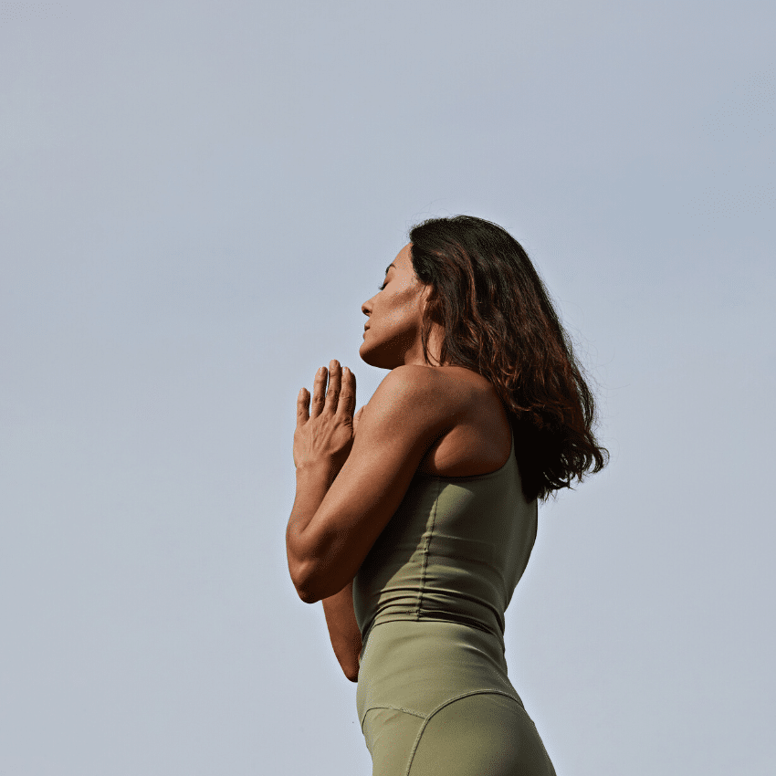 woman with dark hair and a tan in a green yoga outfit with her palms pressed together and eyes closed against a blue sky