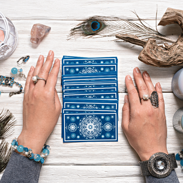 pair of hands covered in rings on a white wooden table on either side of blue tarot cards