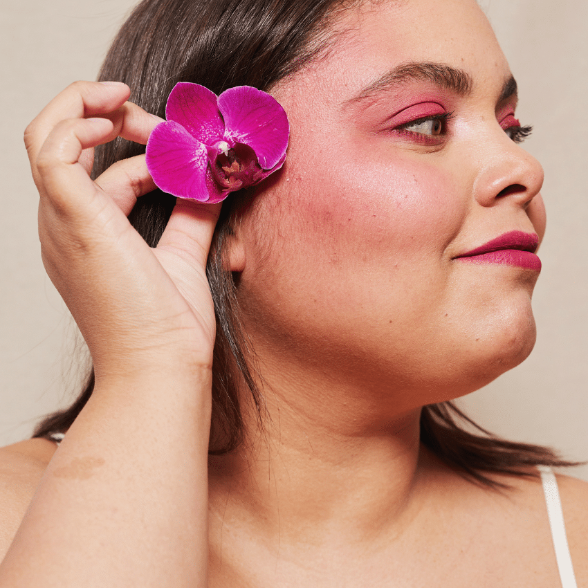 brunette woman with pink lipstick, eyeshadow, and blush holding a pink flower up to her ear
