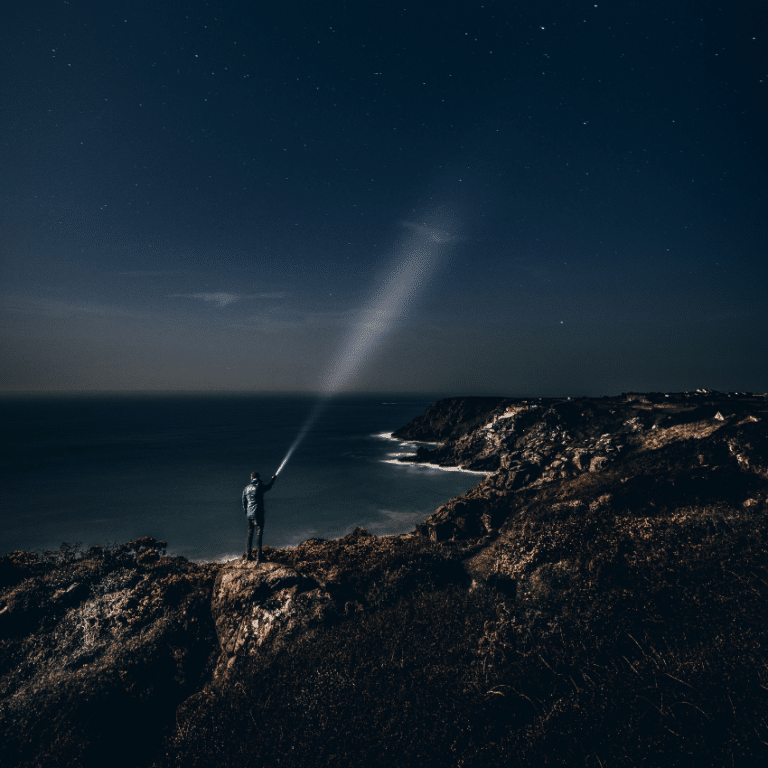 man on brown rock surrounded by water shining a flashlight up to a dark blue sky