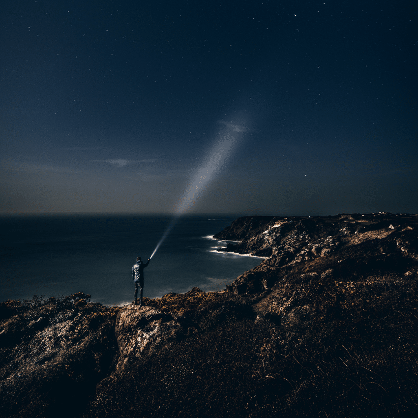 man on brown rock surrounded by water shining a flashlight up to a dark blue sky