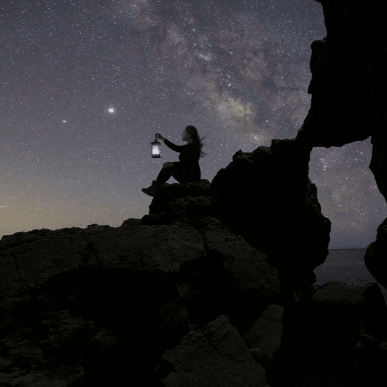 woman sitting on rocks holding up a lantern against a purple starry night