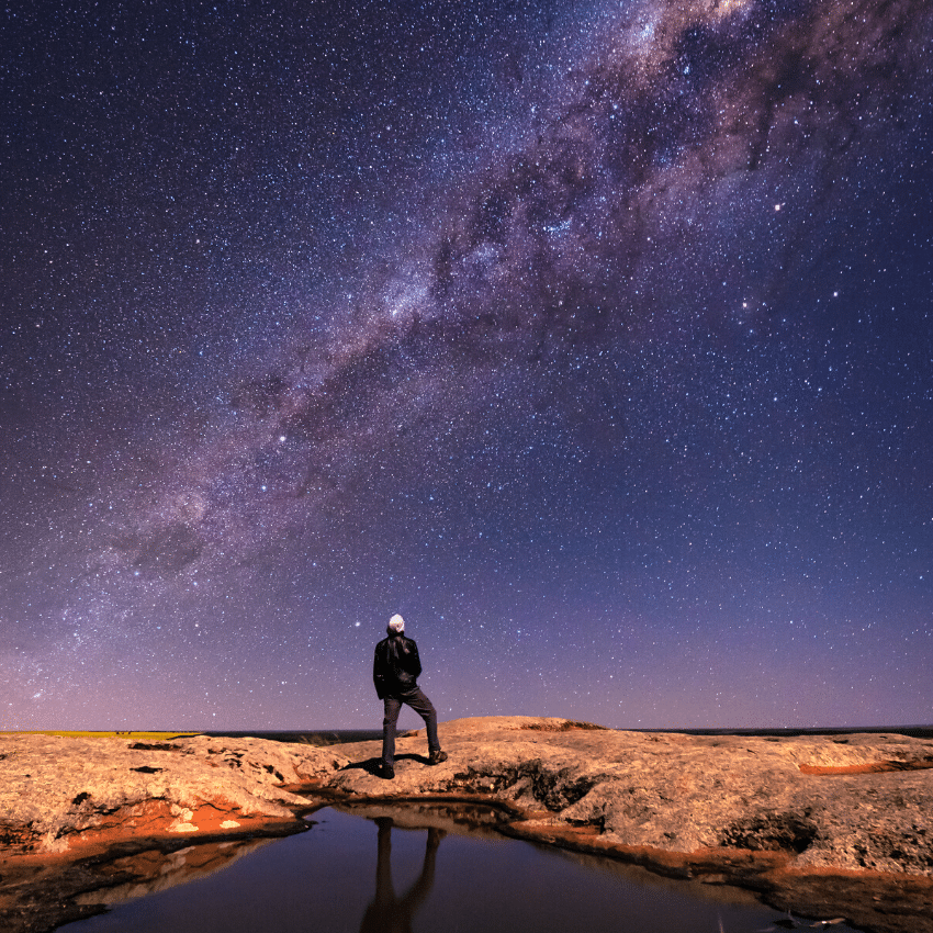 man standing on brown terrain gazing up at a purple starry sky