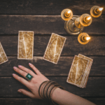 four gold tarot cards on a brown wooden table with a golden candle holder and a woman's hand reaching out with a large emerald ring