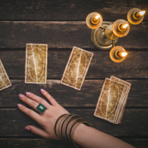 four gold tarot cards on a brown wooden table with a golden candle holder and a woman's hand reaching out with a large emerald ring