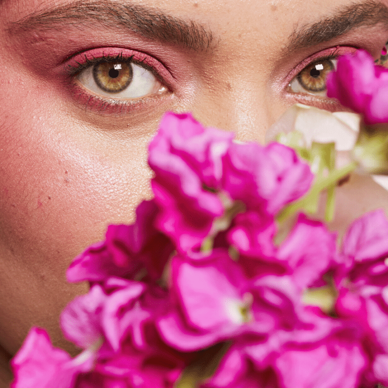 a pair of woman's green eyes, clad with pink eyeshadow, looking over an array of pink flowers
