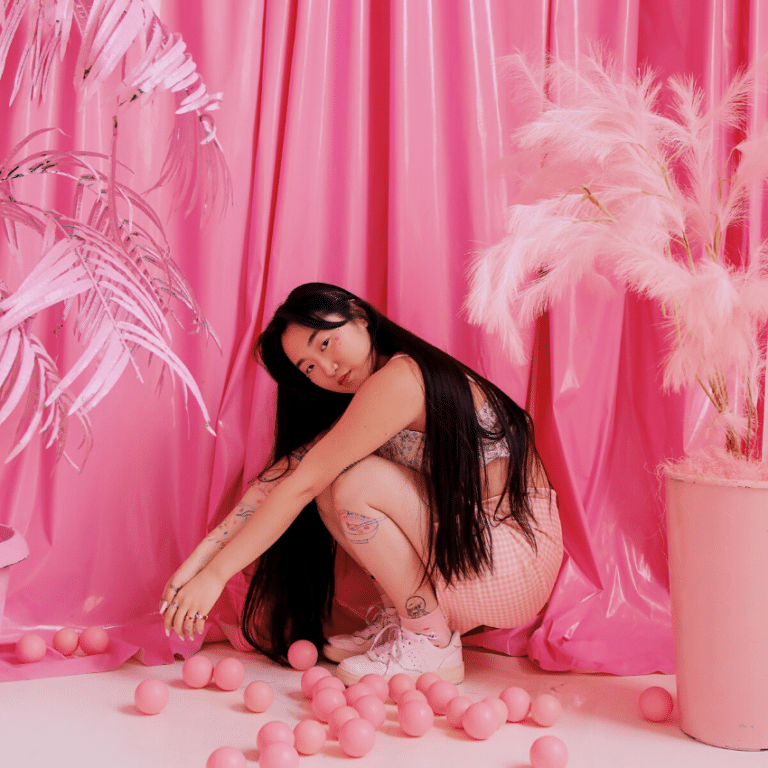 Asian woman in a pink outfit kneeling down surrounded by pink decor and a pink background