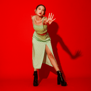 woman in yellow dress and black boots against a bright red background holding out her hand