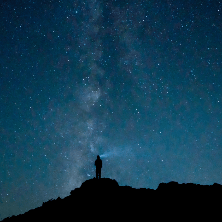 person standing on a large hill starring up at a blue starry sky
