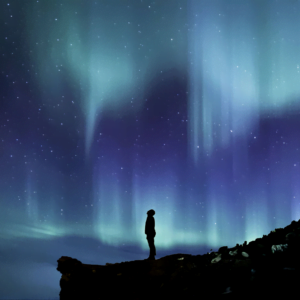 man standing on the edge of a cliff gazing up at a blue and green starry sky