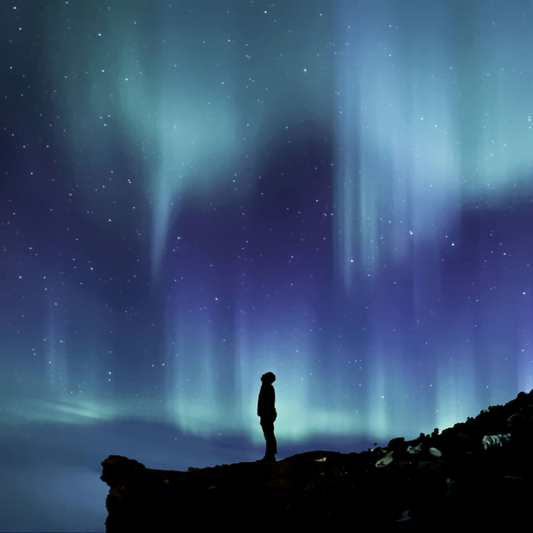 man standing on the edge of a cliff gazing up at a blue and green starry sky