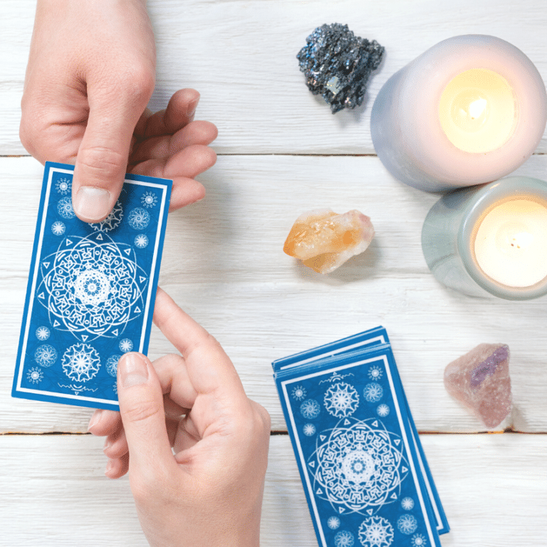 two hands holding onto a blue tarot card over a white wooden table filled with candles, crystals, and a pile of blue tarot cards