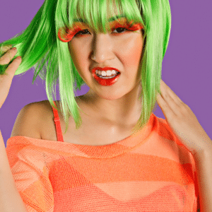 asian woman with short green hair, large orange eyelashes, and an orange shirt against a purple backdrop