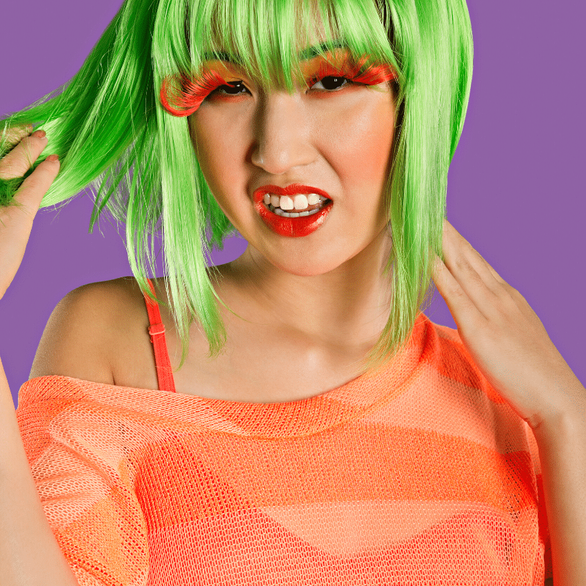 asian woman with short green hair, large orange eyelashes, and an orange shirt against a purple backdrop