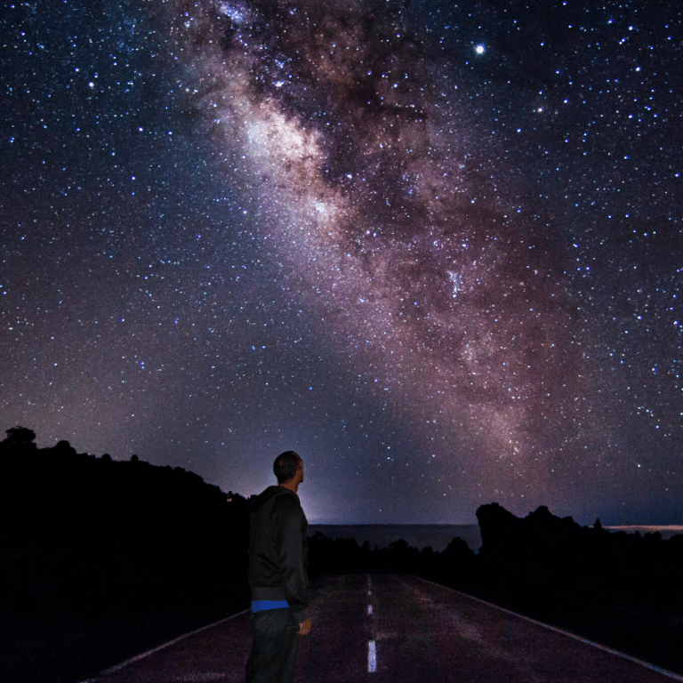 man standing on a road gazing up at a purple starry sky