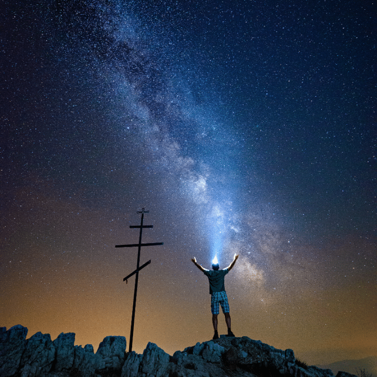 man standing on a cliff with a headlamp on and his hands thrust upward to a starry night sky