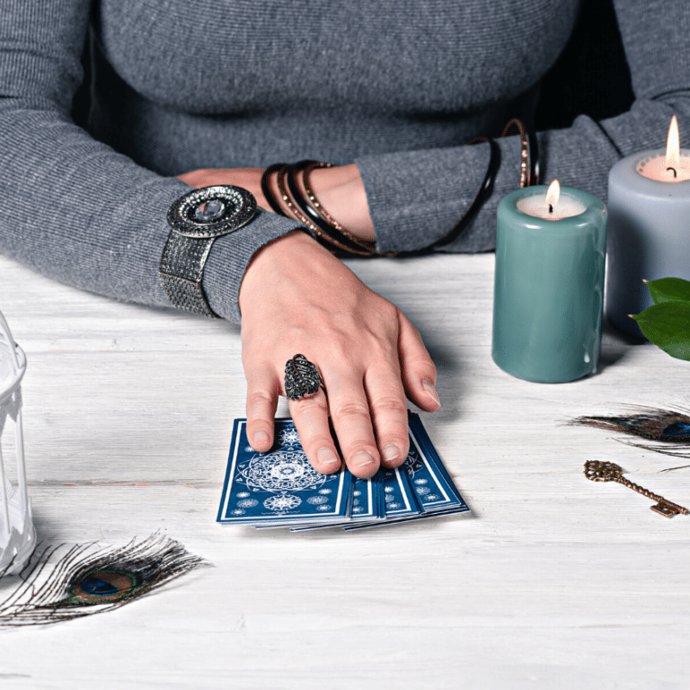 woman in a grey sweater with her hand placed on a pile of blue tarot cards on a white table filled with candles