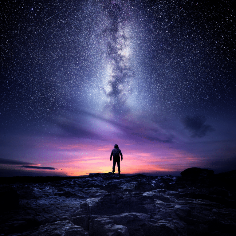 silhouette of a man standing on a ledge of rocks against a pink, purple, and blue starry night