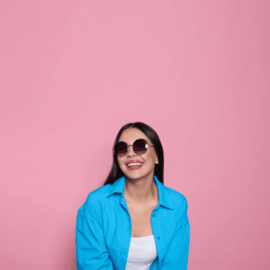 brunette woman in sunglasses and a white and blue shirt smiling against a pink backdrop