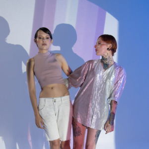 two woman leaning against each other in funky clothes against a blue background