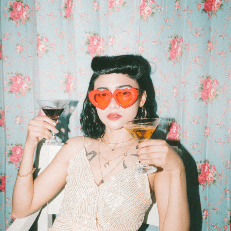 brunette woman wearing red heart-shaped glasses holding up two martini glasses against a backdrop of floral wallpaper