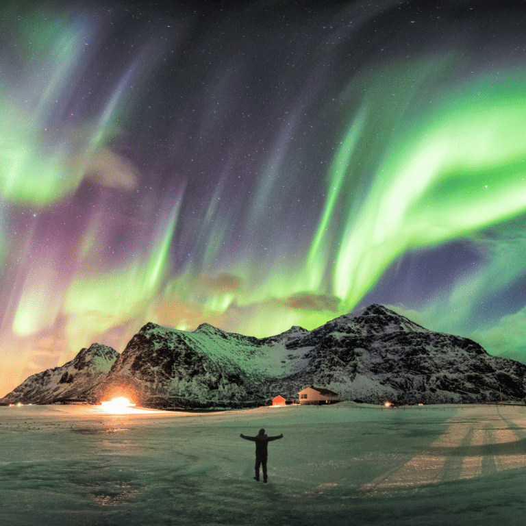 man with his arms outstretched in front of snowy mountains and the northern lights in the night sky