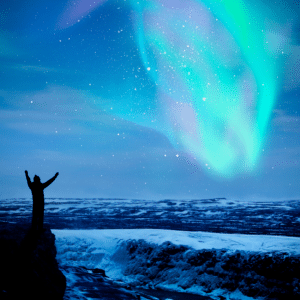 silhouette of a person standing on a cliff with their hands outstretched to the northern lights in the sky