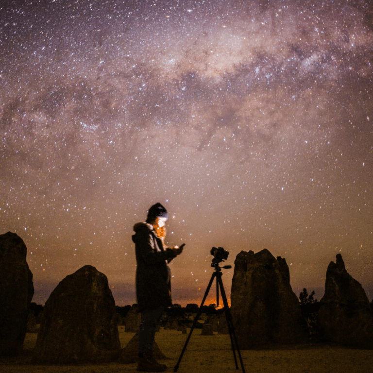 man in the desert surrounded by rocks below a pink and purple starry night