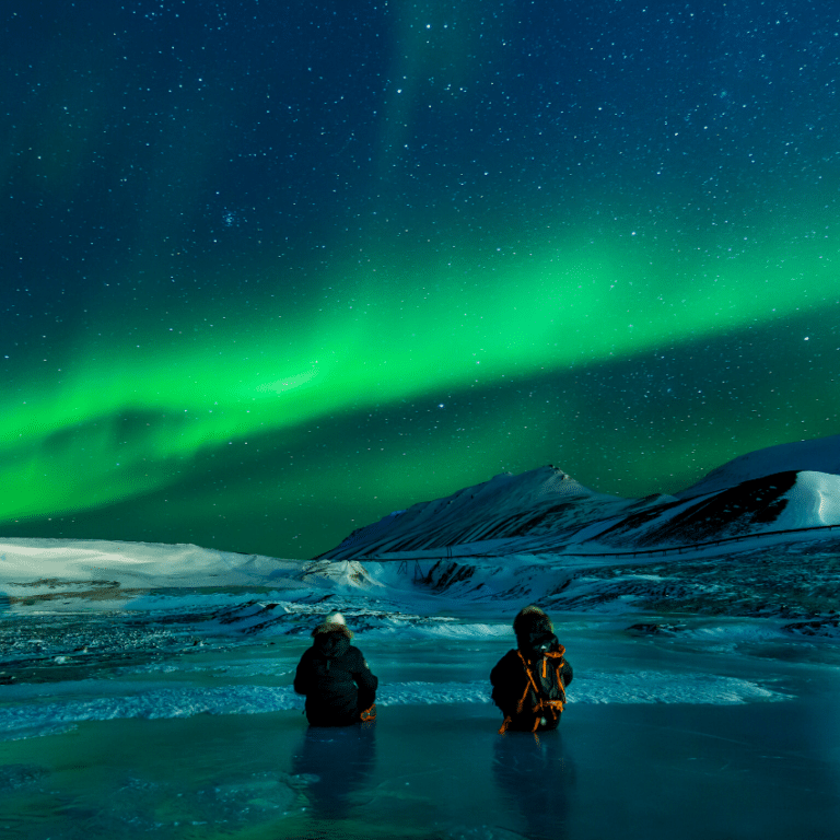 two men in icy waters gazing ahead at snowy mountains and the northern lights in the sky
