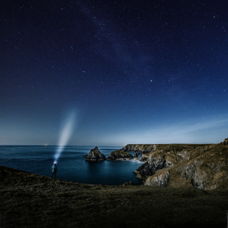 man standing on the edge of a cliff with a shining light against a blue starry night