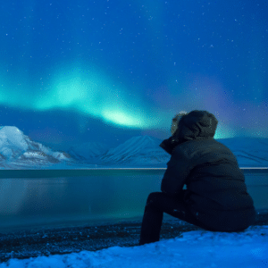 person gazing ahead at the northern lights above snowy mountains