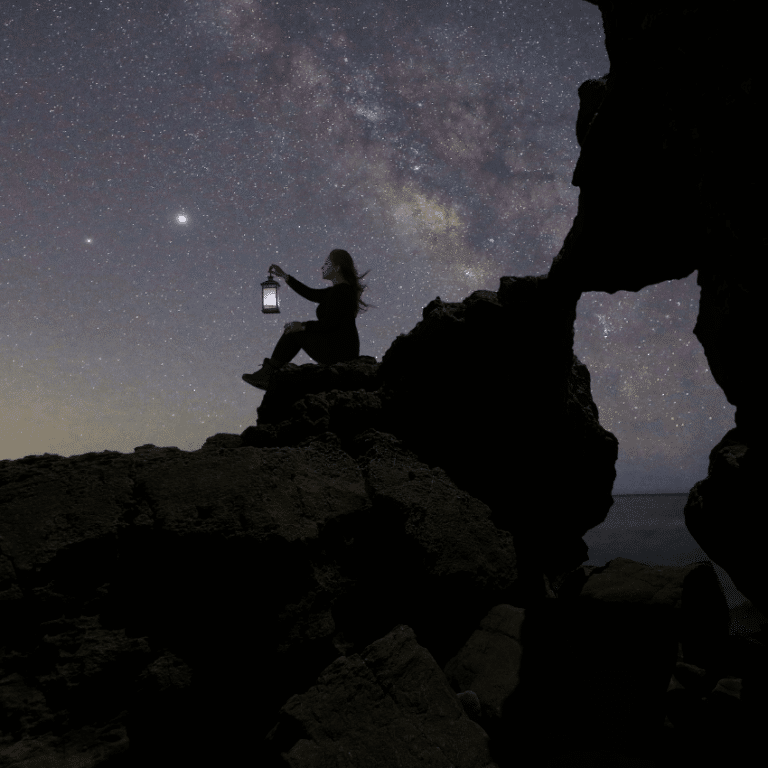 woman sitting on a rock with a lantern gazing at a purple starry night