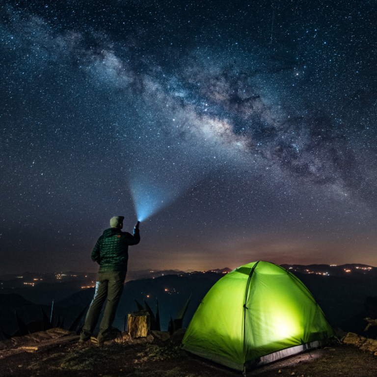 man standing outside of his green tent shining a flashlight up to a starry sky