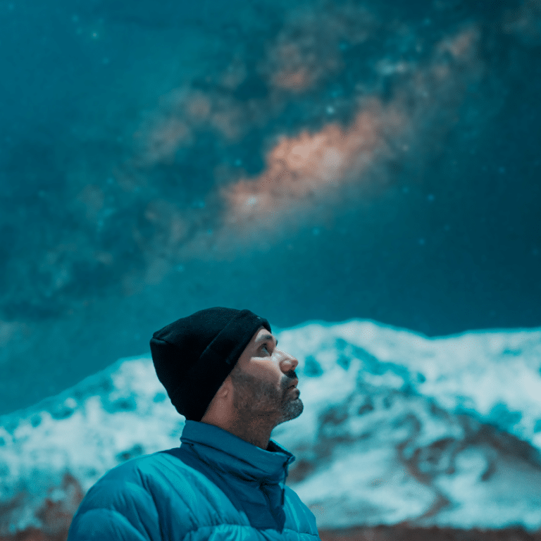 man in a black toque and blue jacket standing in front of a snowy mountain gazing at a blue starry sky