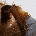 A woman leans down into a field of wheat at sunset.