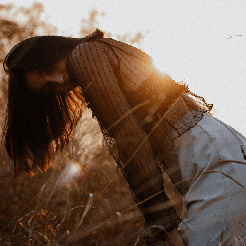 A woman leans down into a field of wheat at sunset.