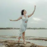 A woman stands on a beach, reaching up into the air to encompass the energy of Aquarius season.