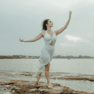 A woman stands on a beach, reaching up into the air to encompass the energy of Aquarius season.